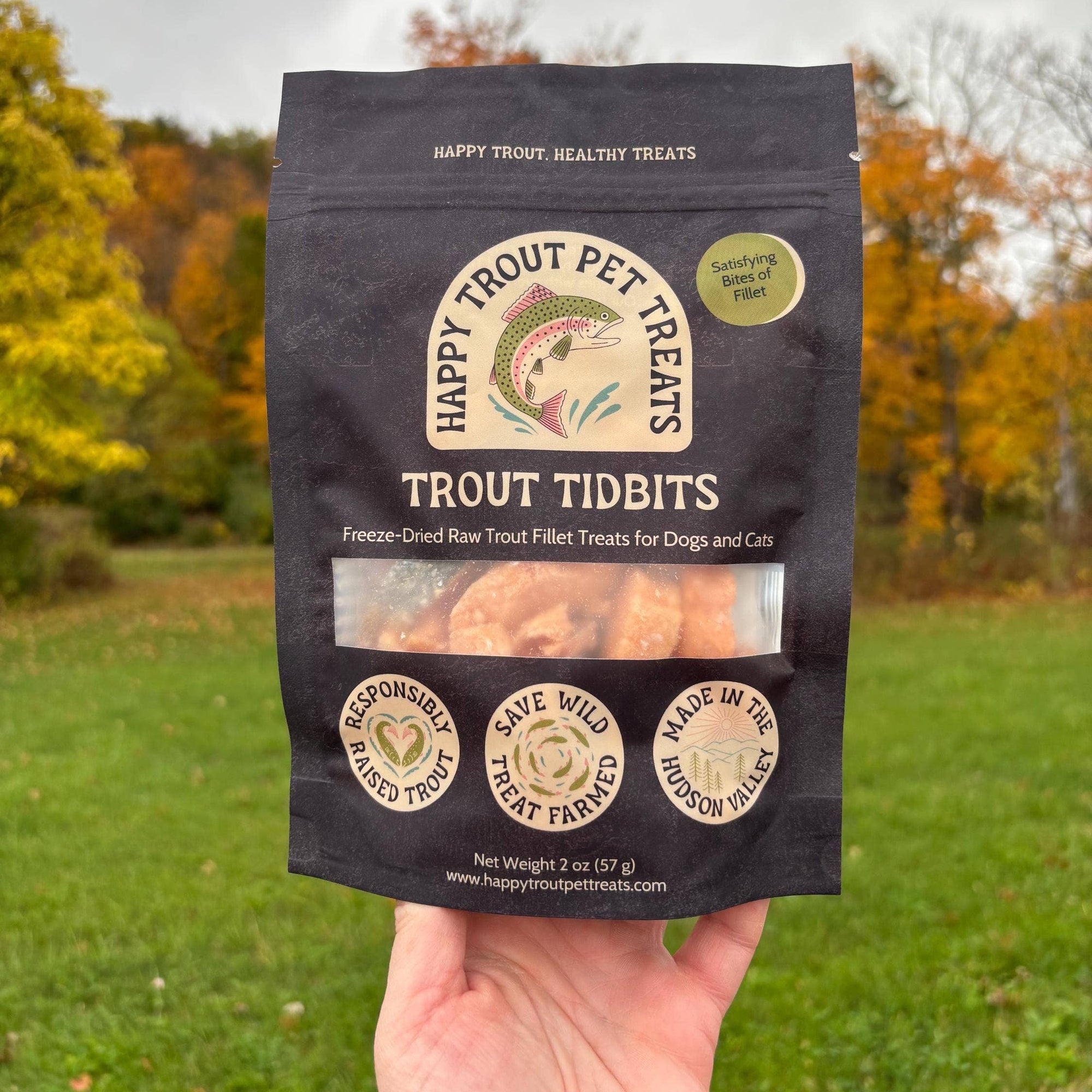 Hand holding a package of Happy Trout Pet Treats Trout Tidbits in an outdoor setting with trees in the background.