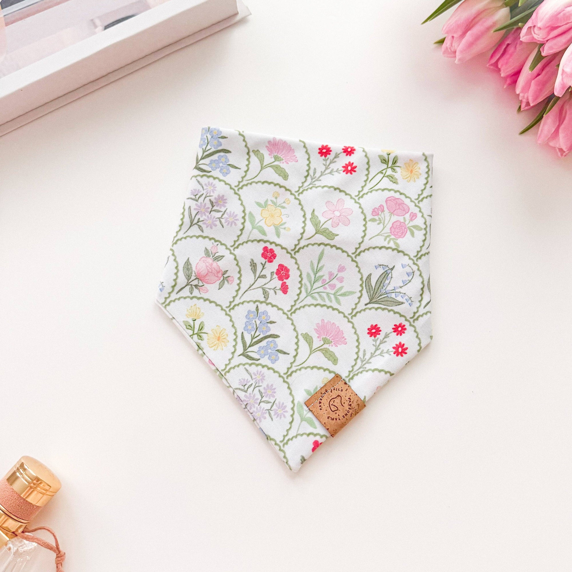 Floral patterned bandana on a white surface with a bottle and flowers in the background