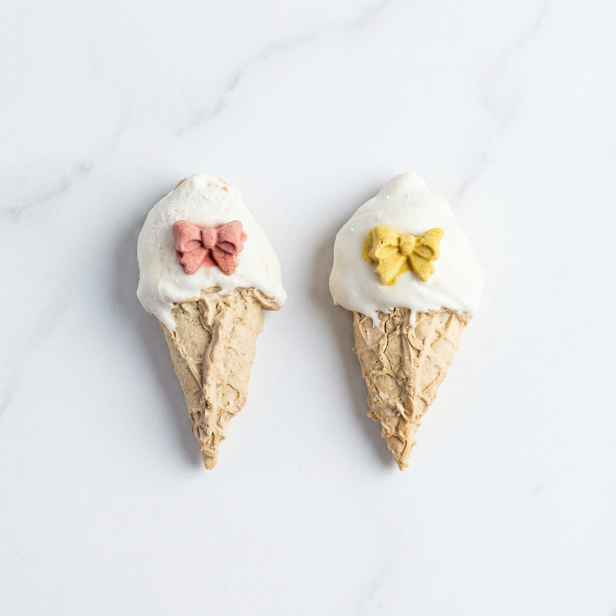 Two ice cream cone-shaped cookies with decorative bows on a white marble surface