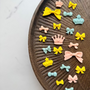 A collection of small decorative bows and crowns in pastel yellow, light blue, and pale pink, arranged on a dark brown wooden tray against a light marble background.
