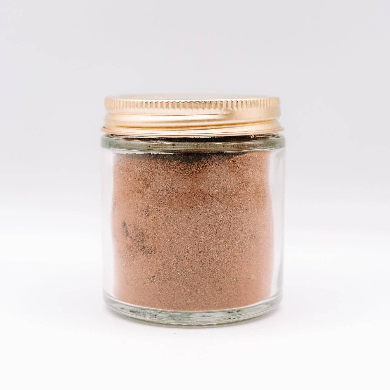 Glass jar with gold lid containing a brown powder on a white background