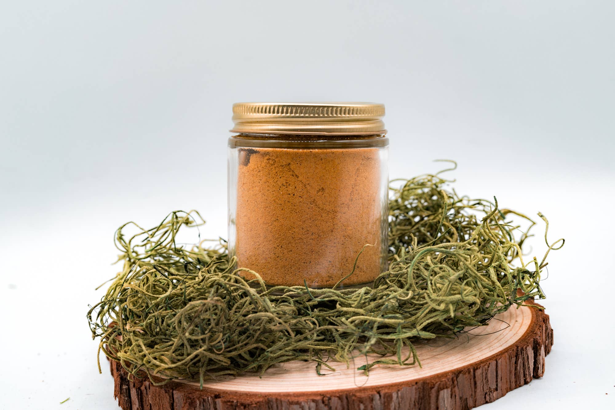 Jar of brown powder with a gold lid on a wooden platform with greenery against a white background